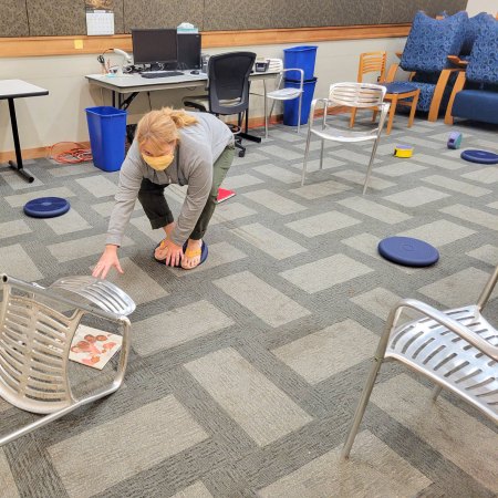 Woman balancing on a pillow trying to pickup a book under a chair.