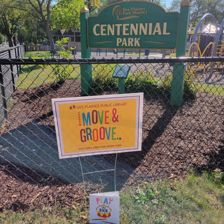 Move and Groove multicolor sign with against fence with Centennial Park sign behind and the book Play that Book by Jessica Young and Daniel Wiseman resting against the sign.