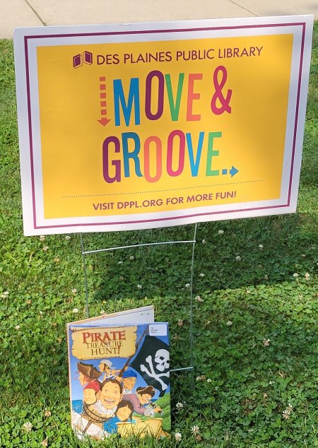 Yellow lawn sign with Move and Groove in multiple colors with the book pirate treasure hunt leaning against it