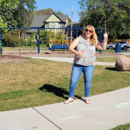 Blonde haired woman in jeans and t-shirt standing on a sidewalk at the park playing air guitar